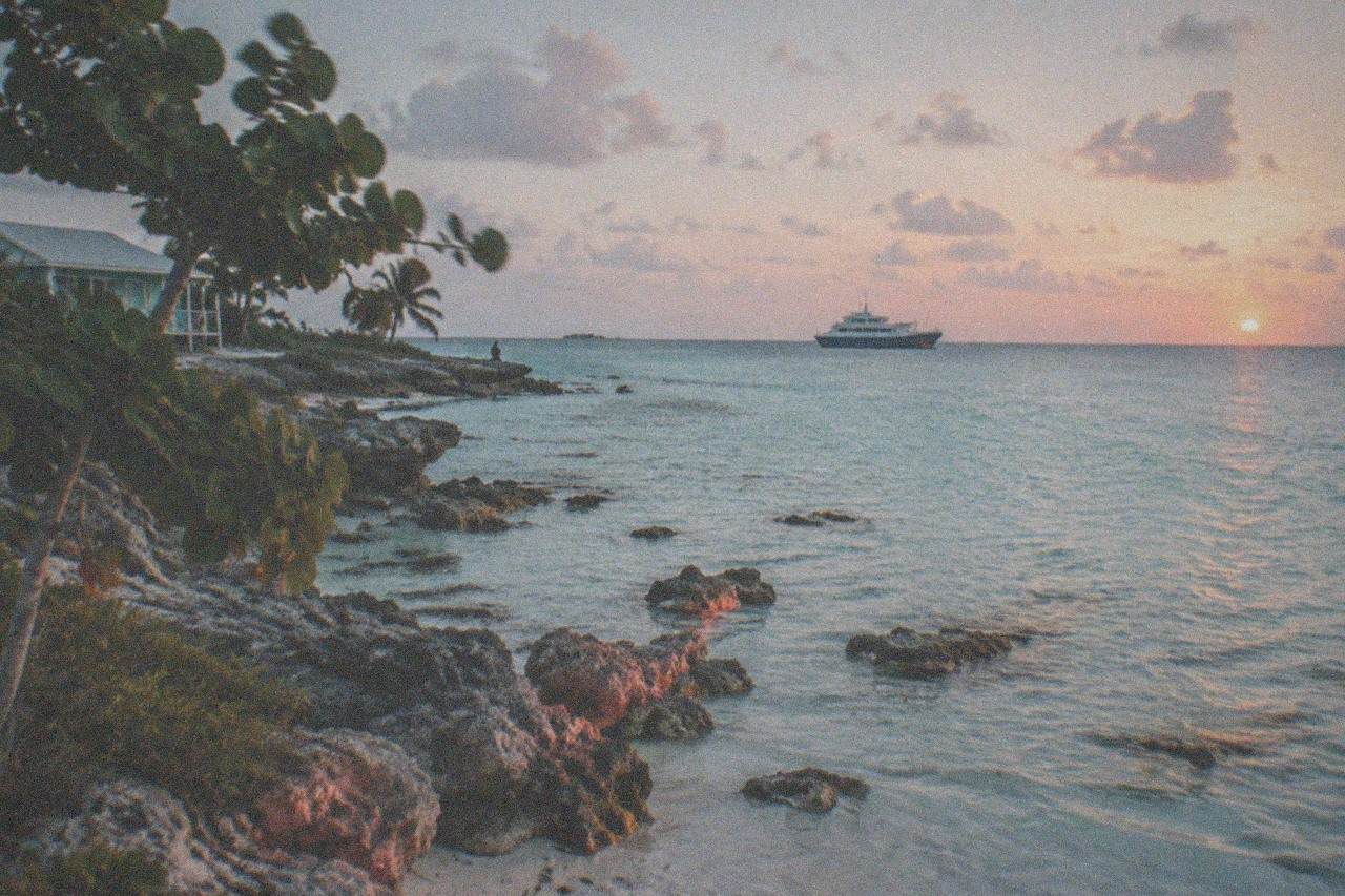 Turquoise shallows along a small Bahamian island shoreline with a distant ferry at anchor