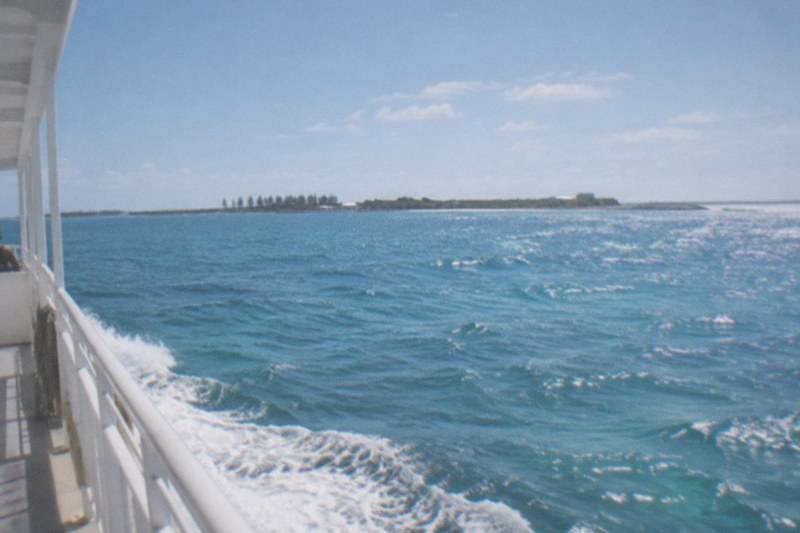 A ferry traveling across calm open sea toward flat island outlines barely visible on the horizon, bright sunlight on the water, no people, no logos, no readable text.