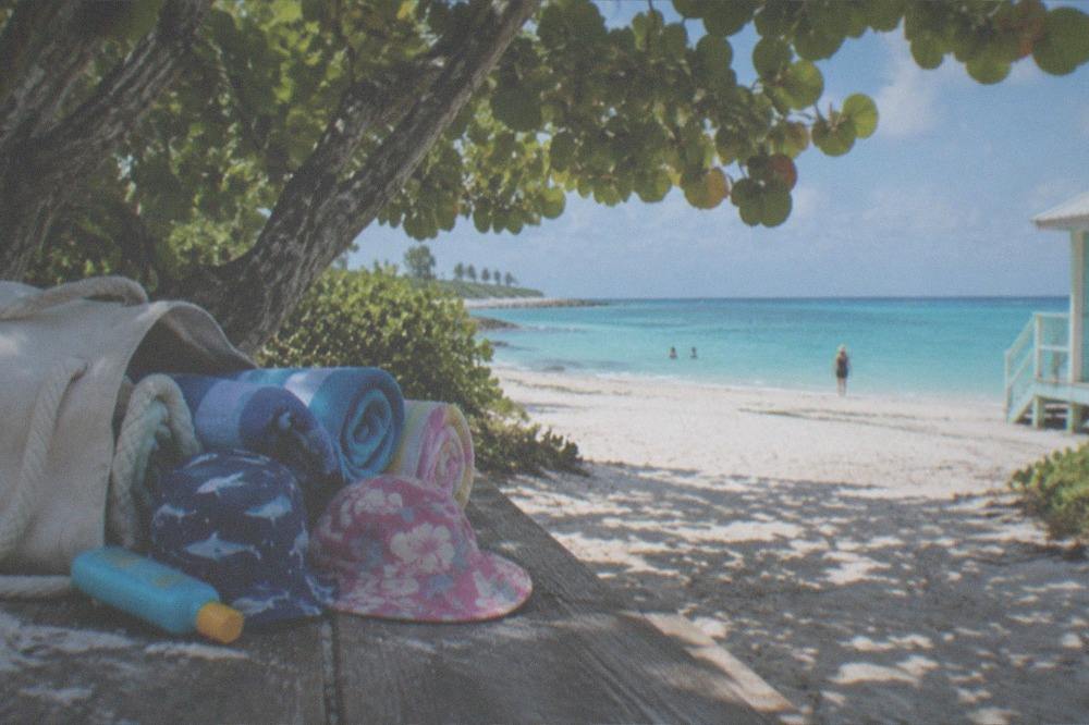 A small beach bag with sunscreen, hats, and folded towels on a wooden bench