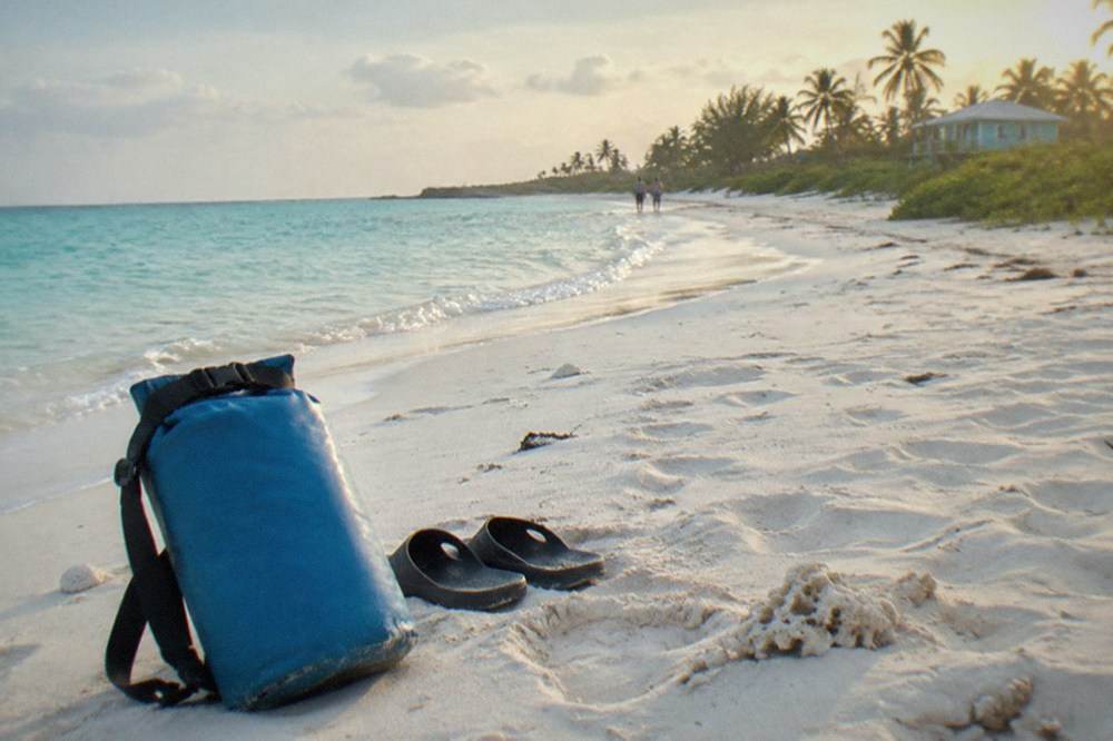A dry bag and sandals resting on a sunlit beach beside calm turquoise water
