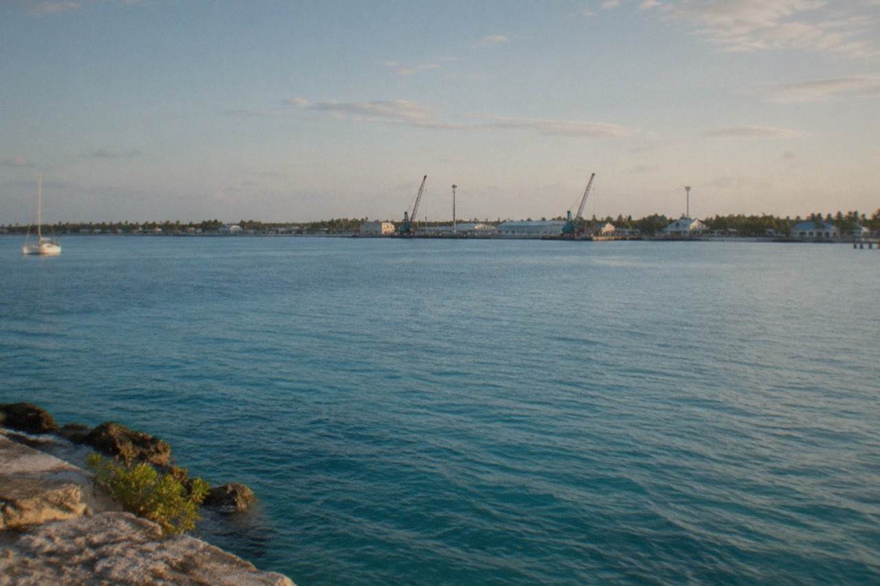 A wide view of a ferry crossing open Atlantic water toward a distant low green coastline, small harbor structures just visible along the shore, no people, no logos, no readable text.